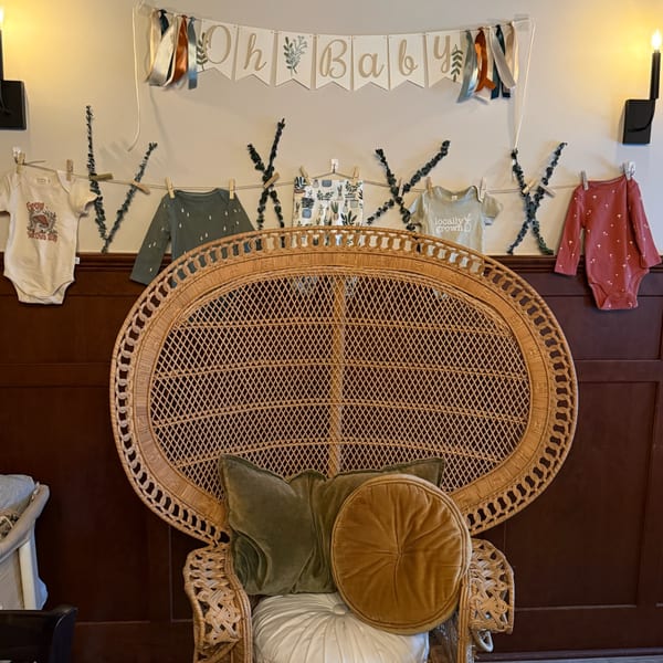 A wicker peacock chair with pillows sits below a baby shower banner and clothesline of baby clothes.