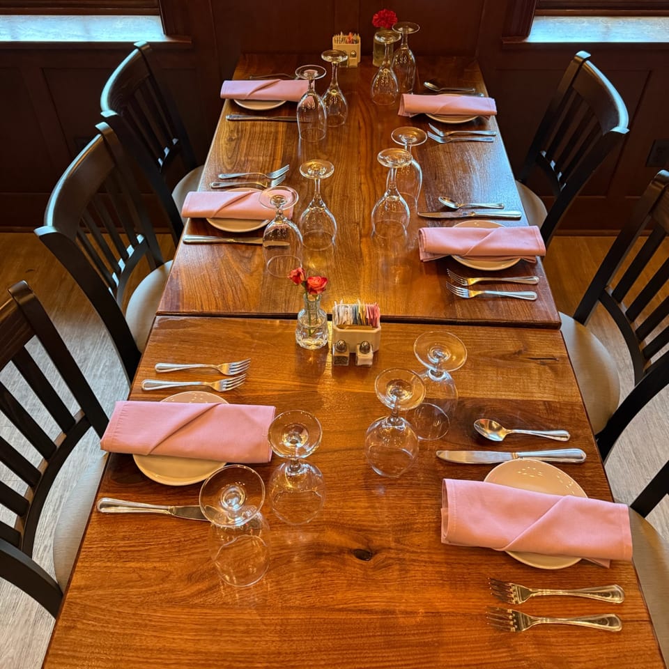 Long wooden restaurant table set with pink napkins, plates, silverware, inverted wine glasses, and small red flower vases.