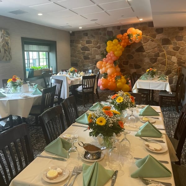 A restaurant dining room set for an event, featuring tables with white cloths, green napkins, and floral centerpieces.