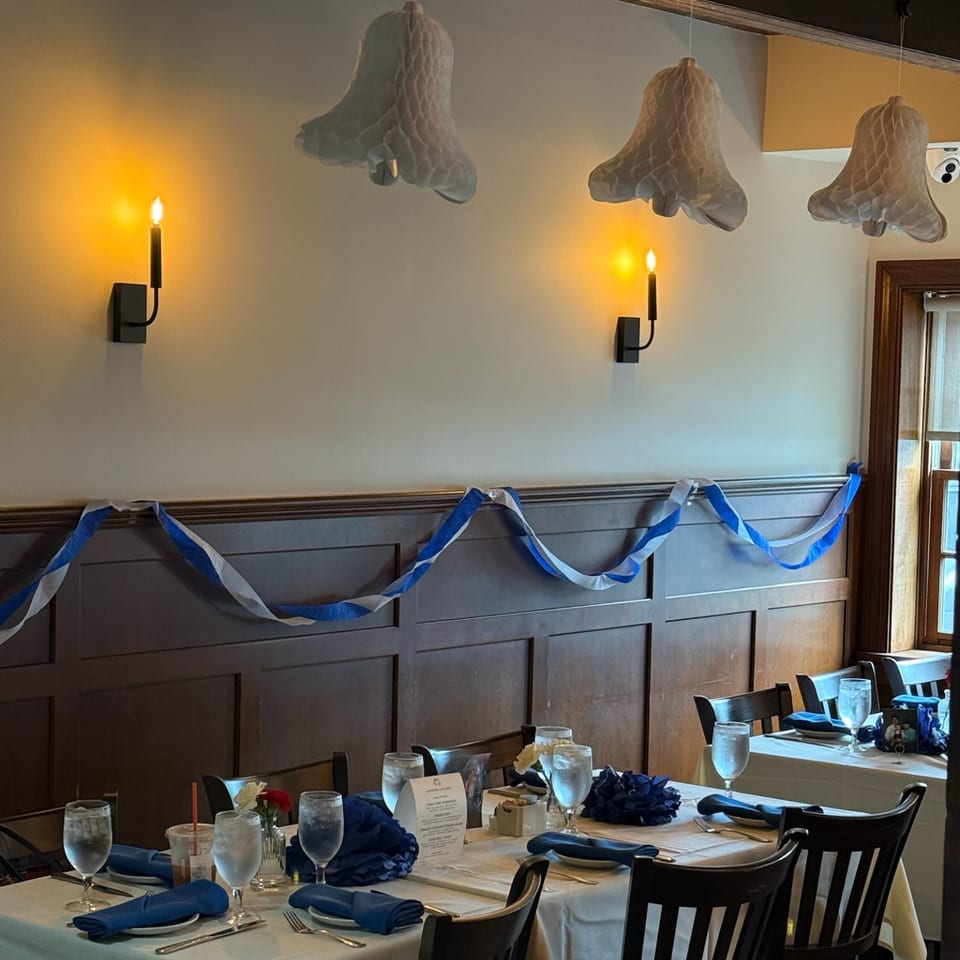 Restaurant dining room decorated with blue-and-white streamers, set tables, hanging floral lamps, and glowing wall sconces.