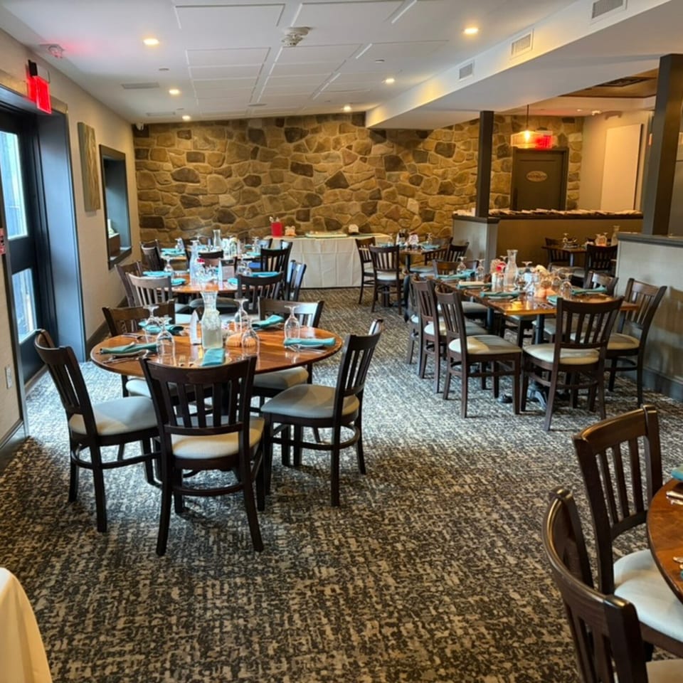 Warmly lit restaurant dining room with stone walls, wooden tables, dark chairs, turquoise napkins, and neatly arranged glassware.