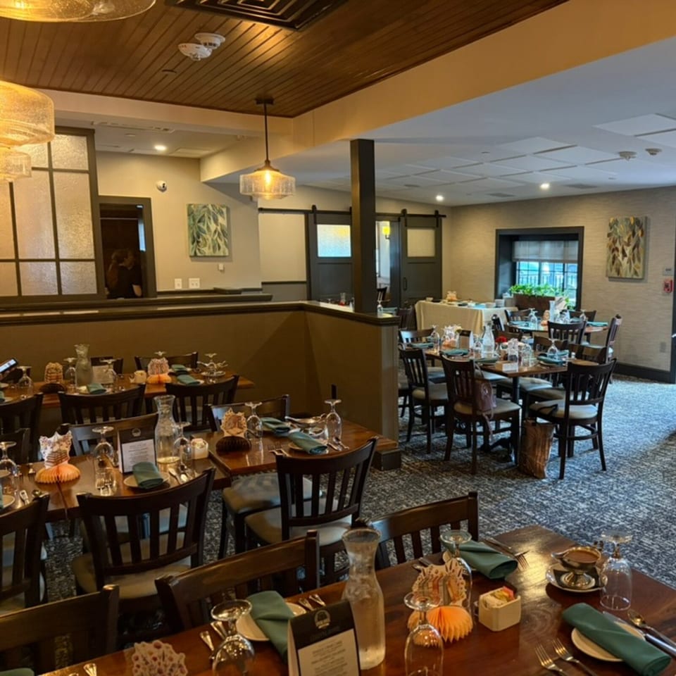 Warmly lit restaurant dining room with wood tables, place settings, dark chairs, pendant lights, and patterned carpeting.