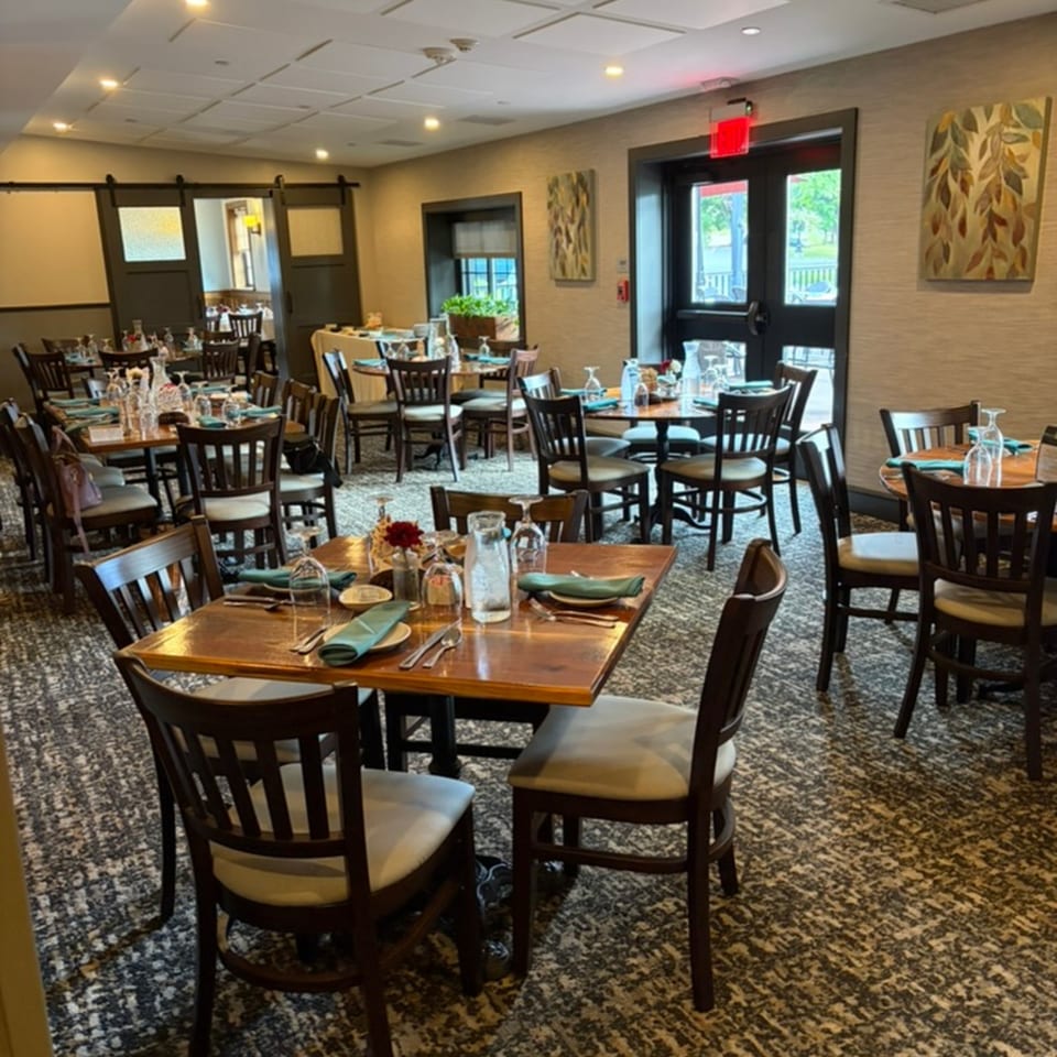 Warmly lit restaurant dining room with wooden tables, set place settings, dark chairs, patterned carpet, and decorative wall art.
